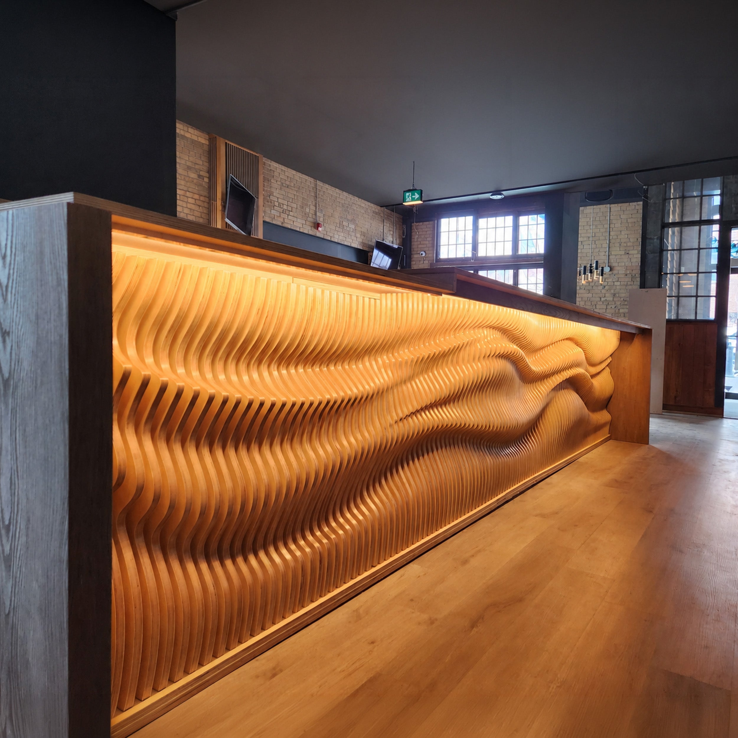 A modern wooden reception desk with a wavy, ribbed front panel illuminated by warm underlight, set in a contemporary office interior with wood flooring and brick walls.