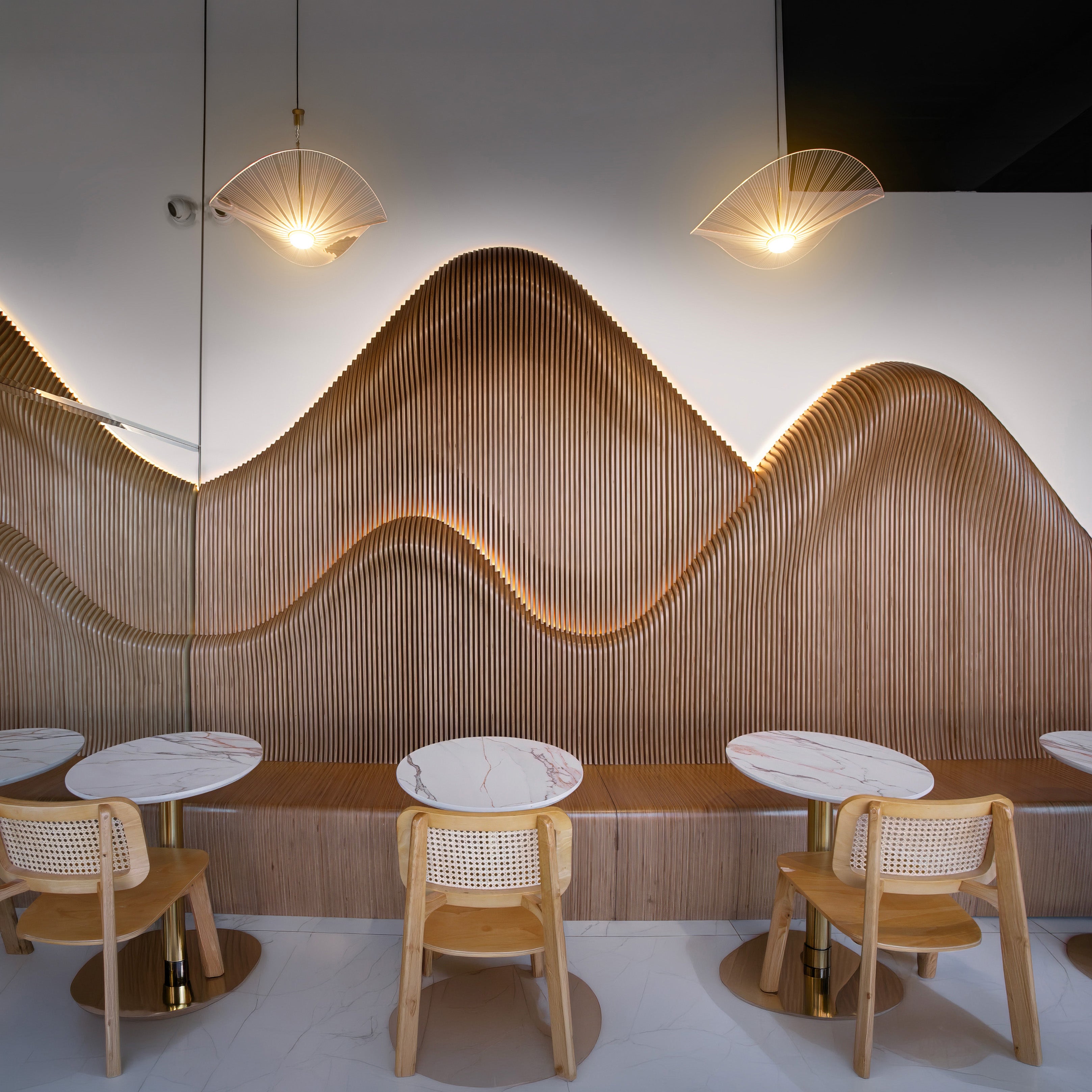 A modern interior seating area with curved wooden slat wall panels, small round marble-top tables, and light wood chairs under two fan-shaped pendant lights.