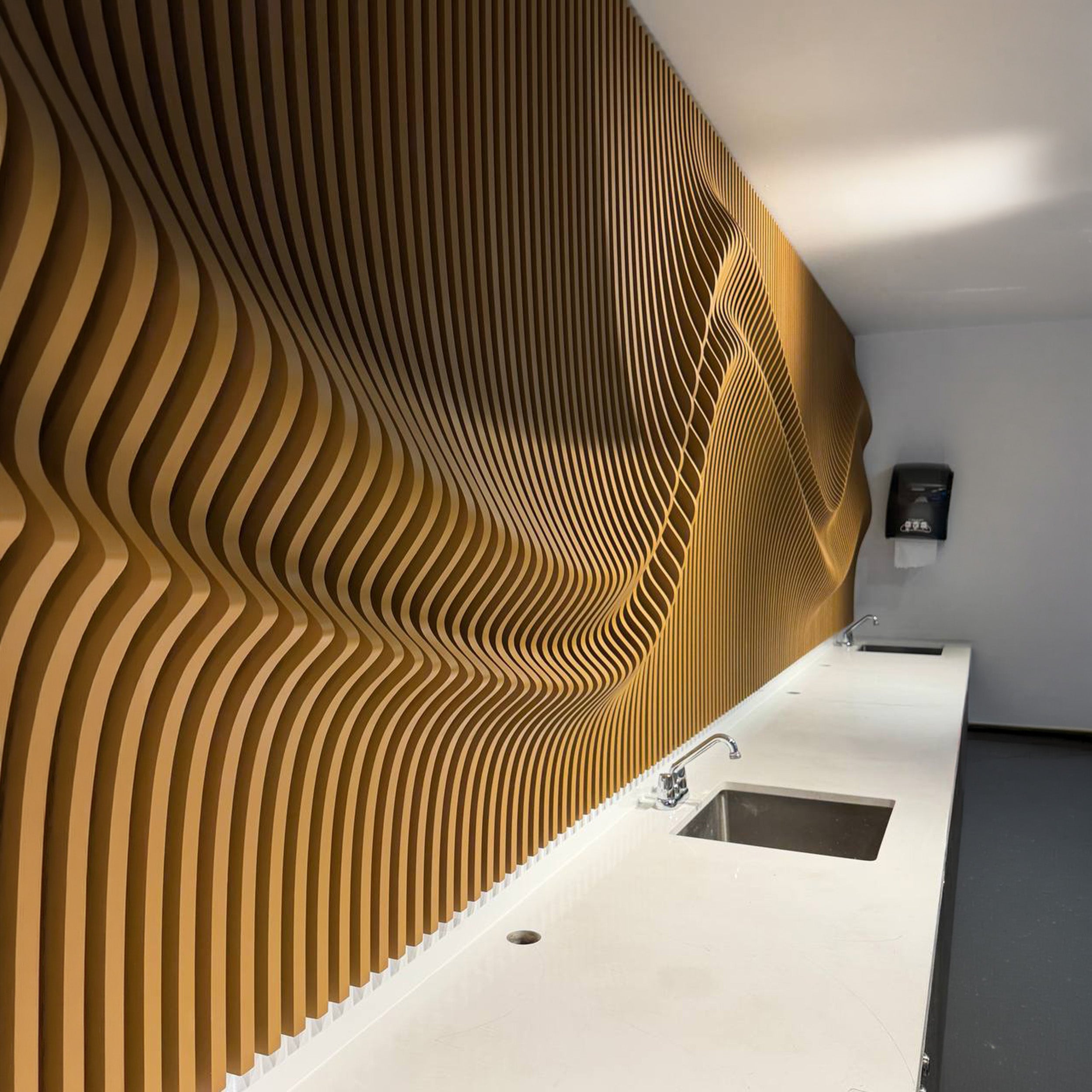 A modern interior space featuring a prominent wavy wooden slat wall in warm brown tones, paired with a long white countertop and sink.