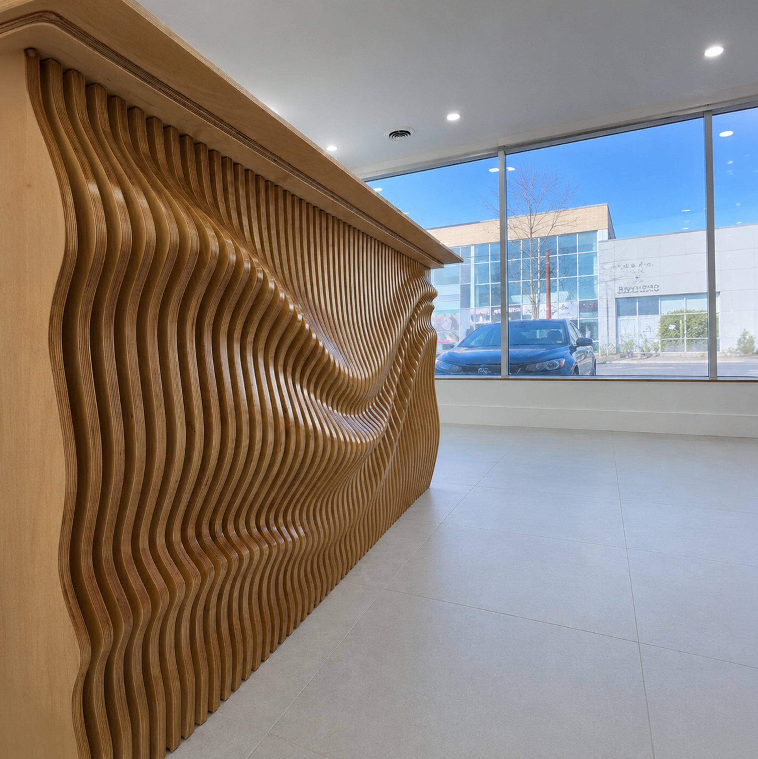 A modern wooden reception desk with a wavy, slatted design, situated in a bright office lobby with large windows showing a blue sky and a car outside.