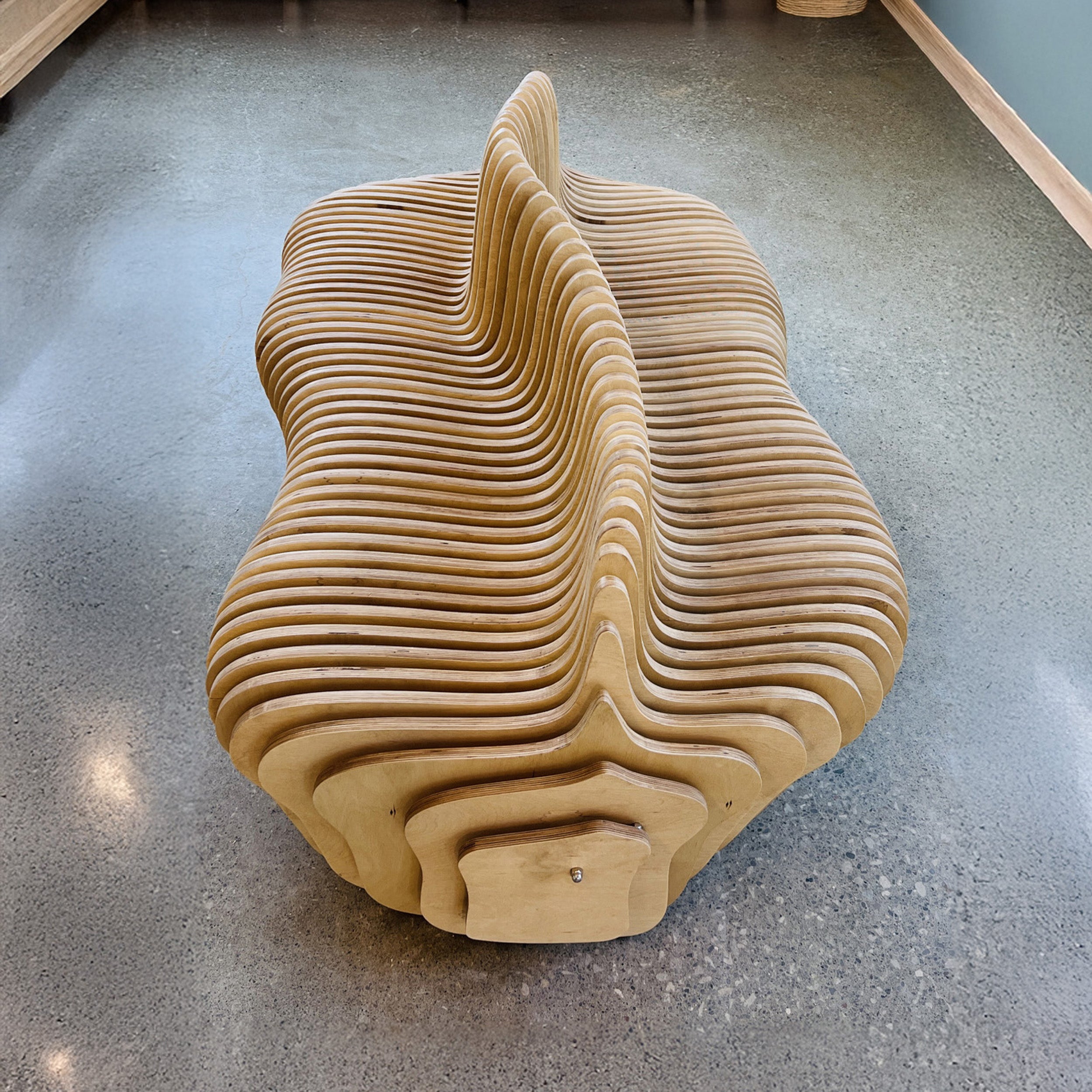 A sculptural, wavy wooden bench made of layered laser-cut wood, shown from a high angle on a polished concrete floor.