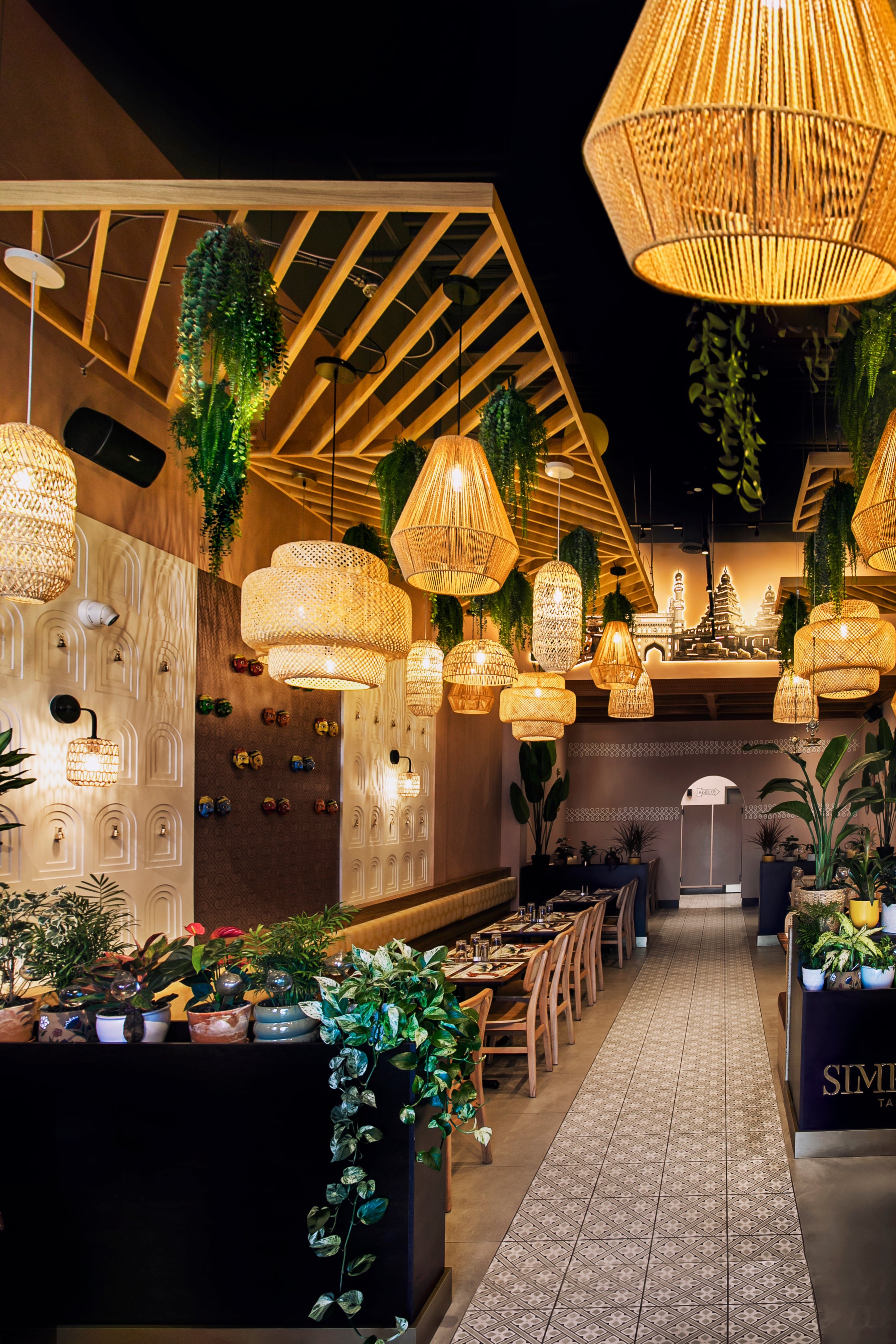 A long dining corridor in a restaurant or cafe with warm woven pendant lights hanging from a high wooden ceiling, green trailing plants, patterned tile flooring, and dark walls.