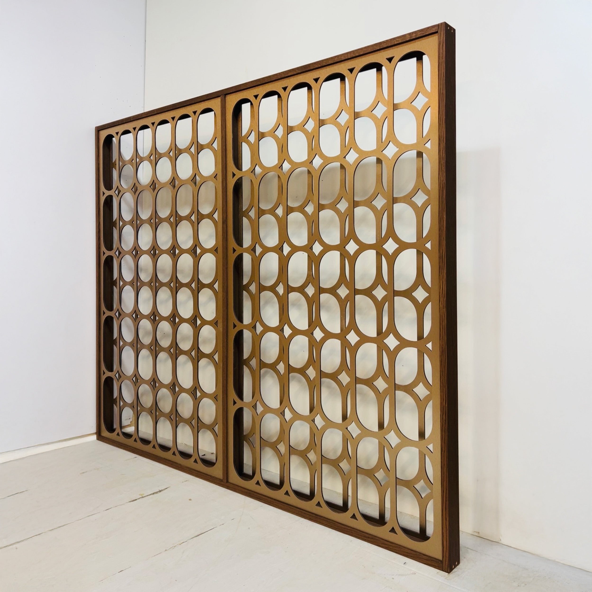 A large rectangular decorative screen panel with an open geometric lattice pattern in a warm metallic gold/bronze finish, leaning against a white wall on a light floor.