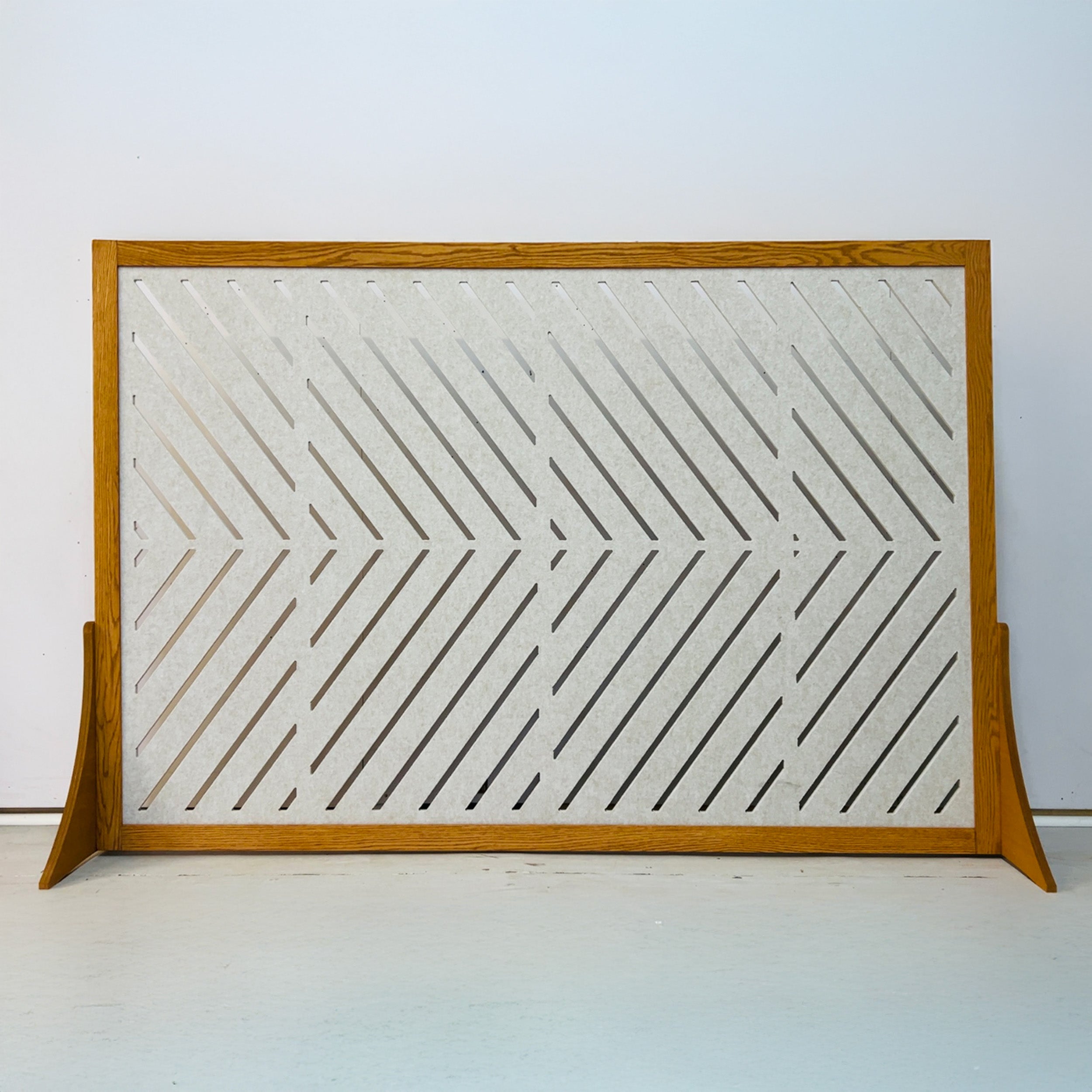 A mid-century modern wooden screen or panel with a warm oak finish, featuring a geometric chevron pattern of diagonal cutouts on a textured off-white panel, supported by two angled feet.