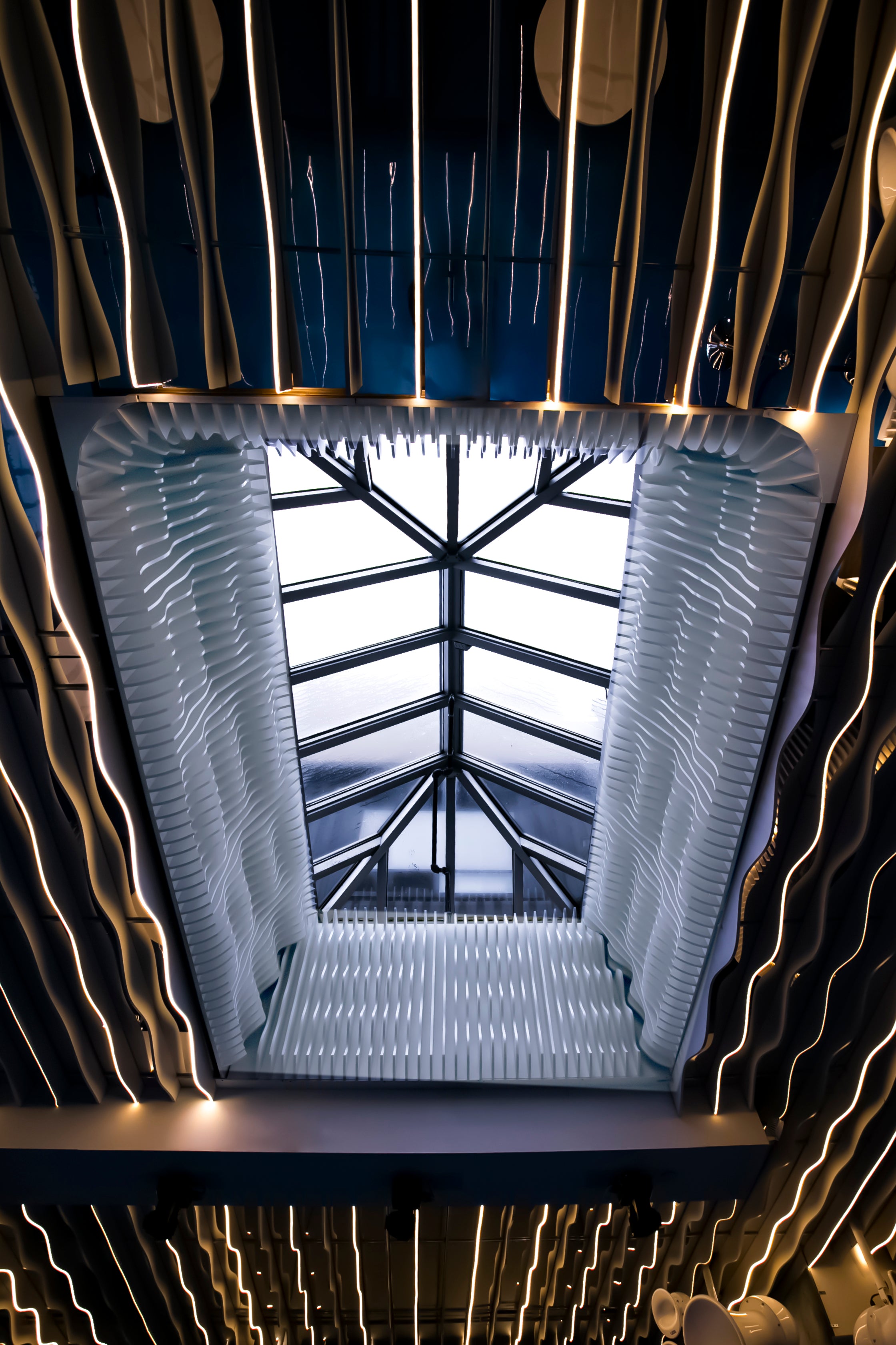 An upward-looking view of a modern architectural skylight with ribbed translucent panels and glowing linear lighting around it.