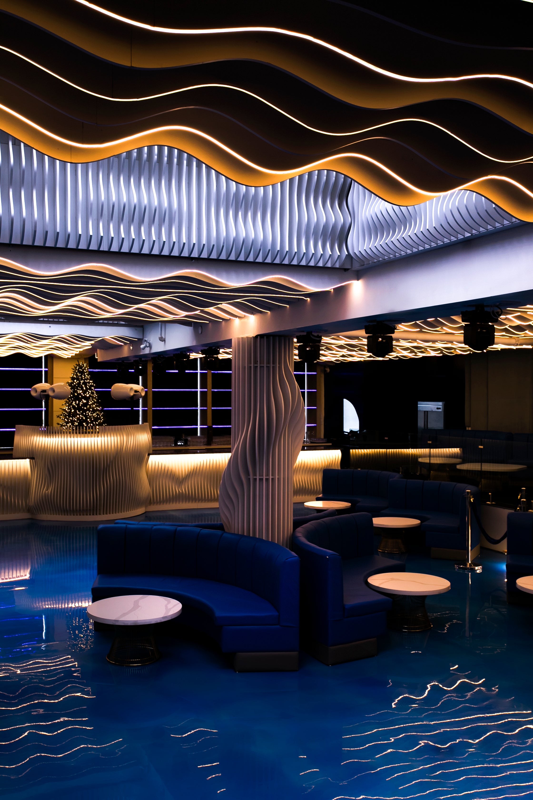 A modern lounge or bar interior with curved blue upholstered seating, small round tables, a wavy slatted ceiling, and blue reflective flooring.