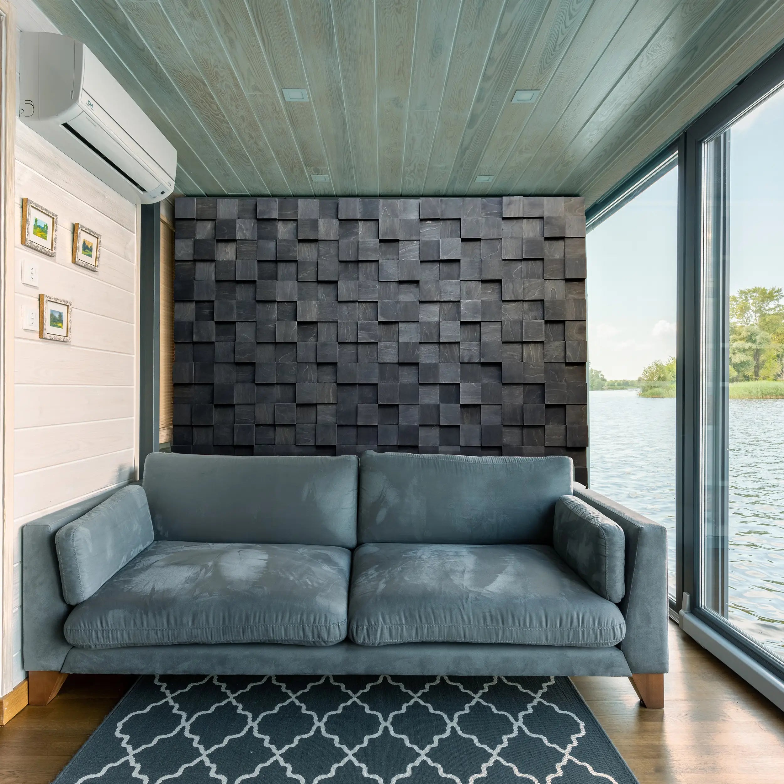 Modern interior with a gray sofa against a textured wall by large windows overlooking water.