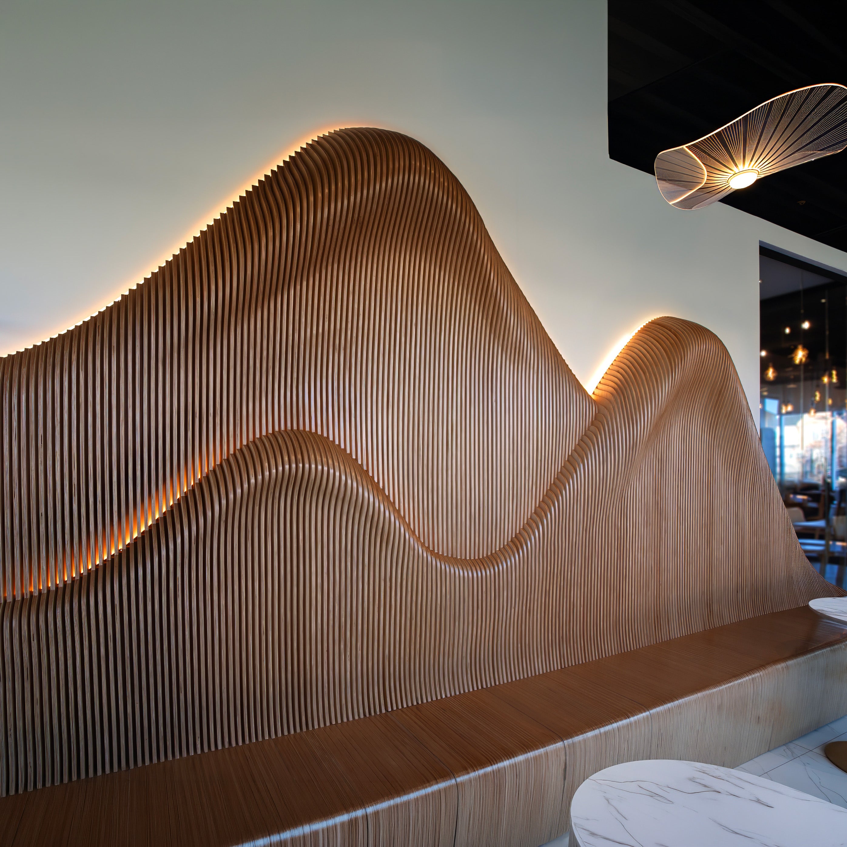 A modern wavy wooden slat wall sculpture or seating feature in a warm wood tone, illuminated by warm under-gaps, set in an interior space with a hanging light fixture and table visible.