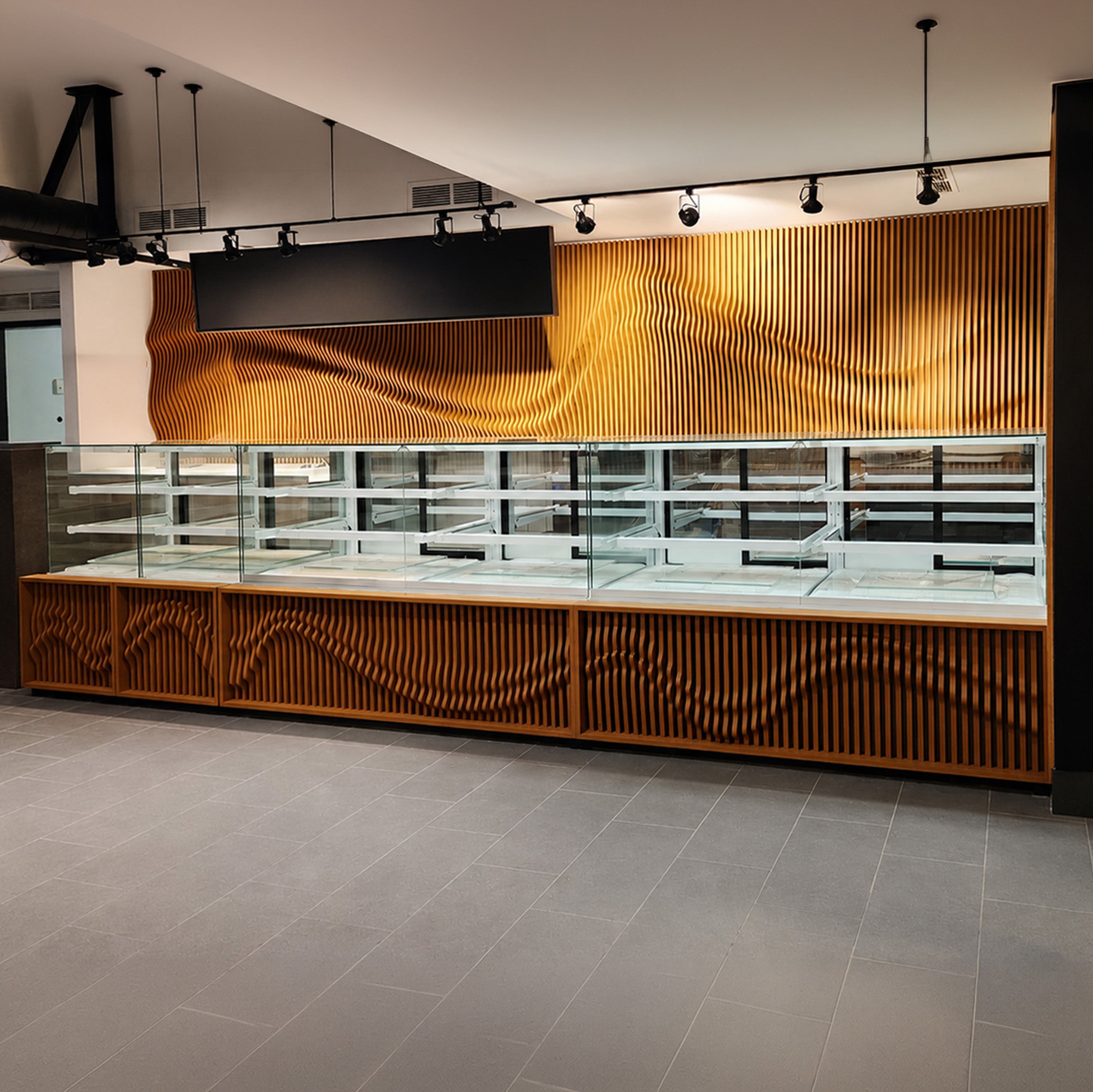 A modern retail display case with glass shelves and wavy slatted wood paneling, set in a clean interior with gray tiled flooring and track lighting.